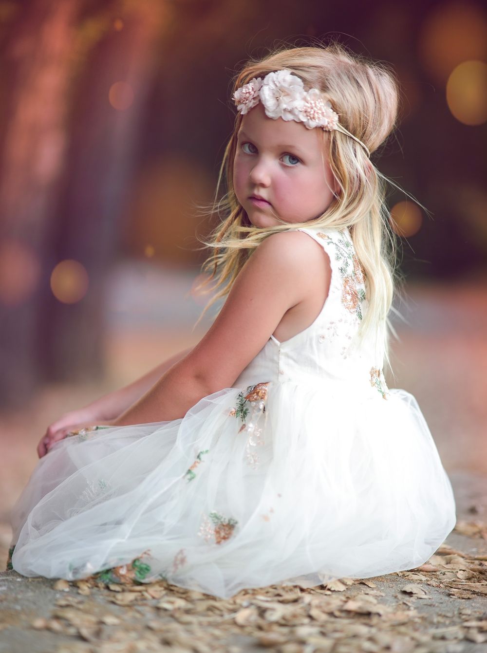 Blonde girl in white floral tulle dress with pink headband sitting outdoors in golden light.