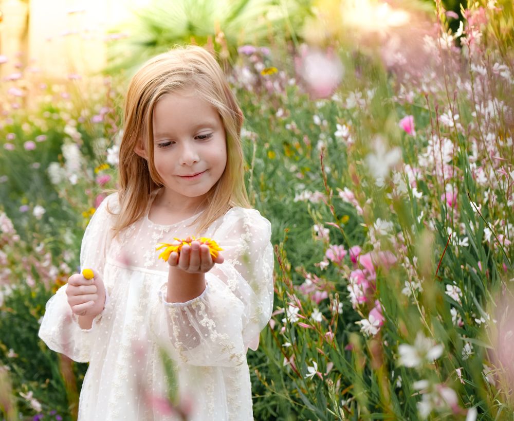 Young blonde girl in white lace dress holding yellow flowers in a wildflower meadow.