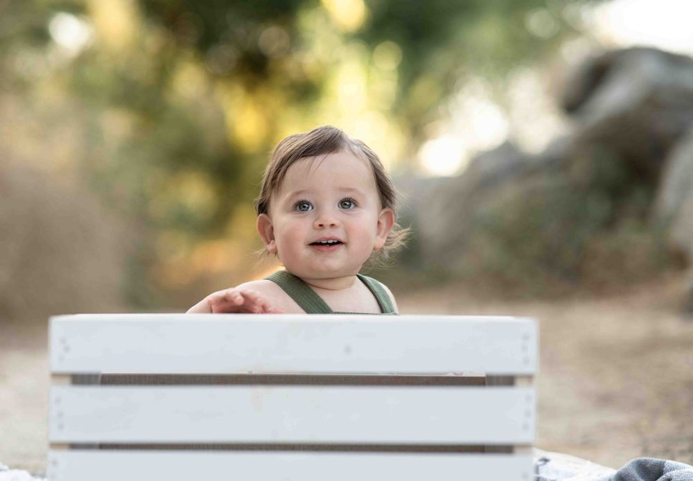 Happy baby peeking over a white wooden crate outdoors in natural light.