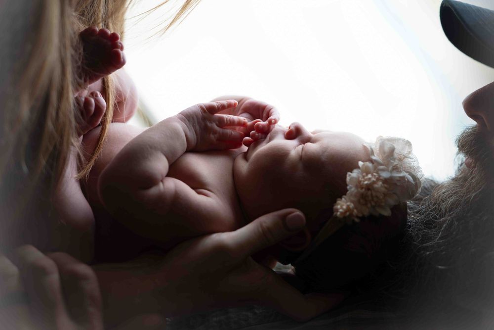 Newborn baby cradled in loving hands with soft natural light and floral headband.
