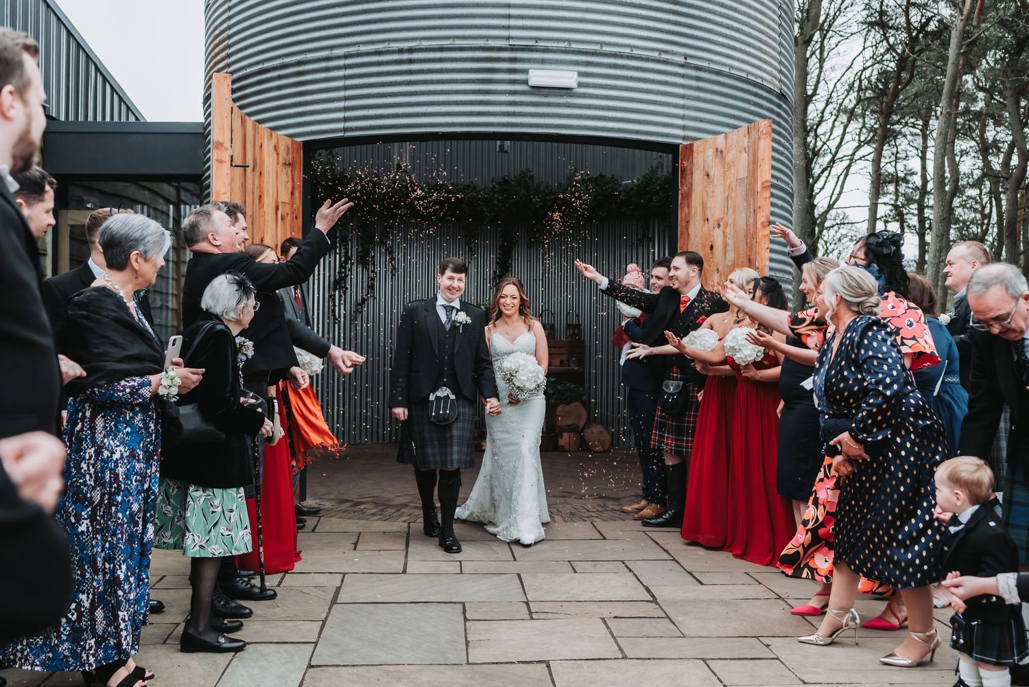 Wedding guests line a stone walkway throwing confetti at a couple exiting a rustic venue.