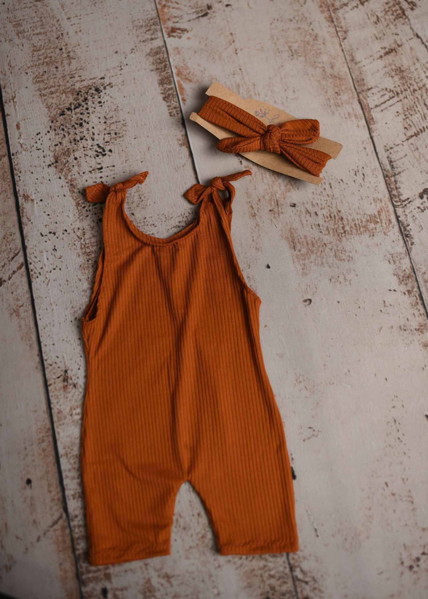 Rust colored ribbed romper with bow straps arranged on distressed wooden background.