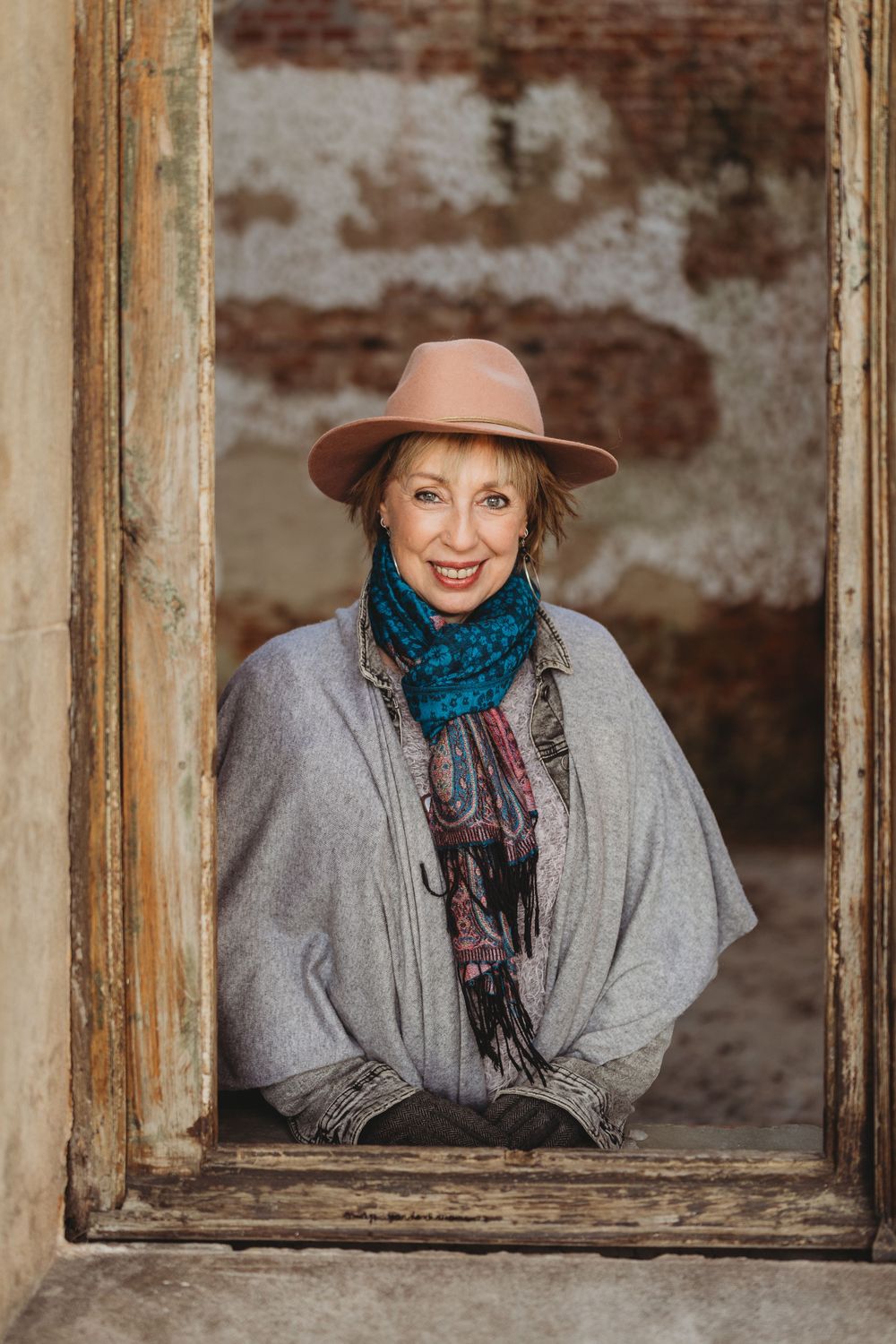 Person in gray wrap and teal scarf smiling through rustic window frame.