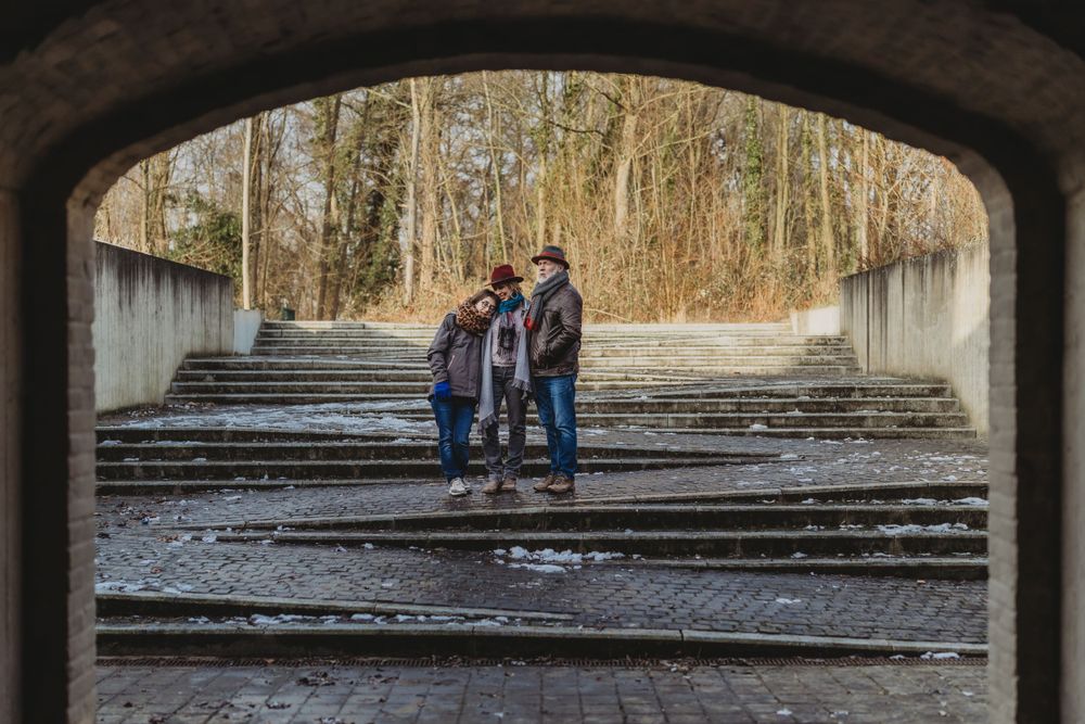 A couple stands together under a concrete archway with steps and trees visible in the background.