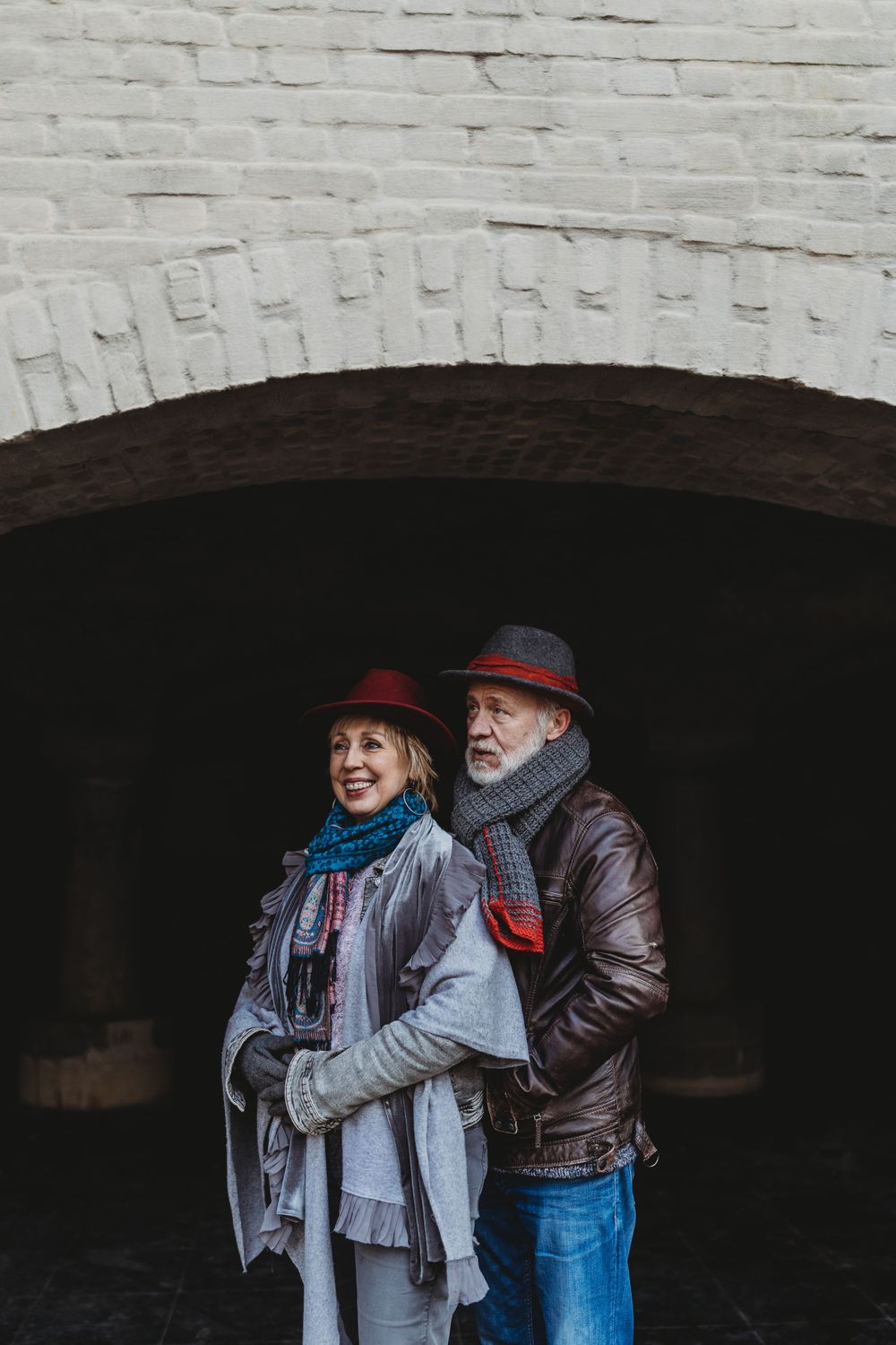 A couple wearing coordinated winter outfits and hats stand close together under a brick archway.