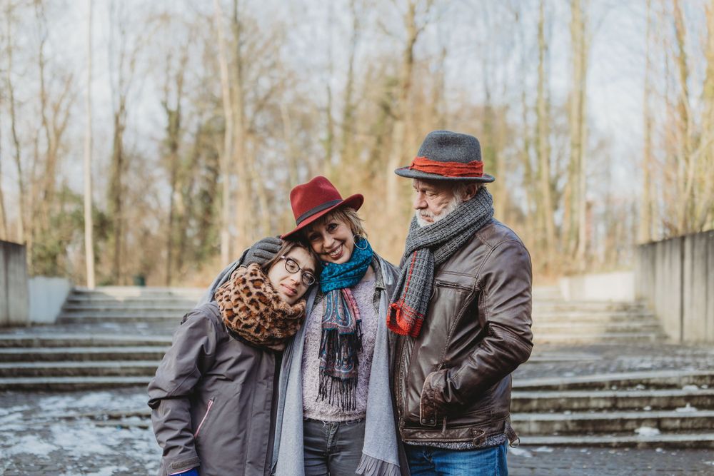 A couple in winter coats and hats embrace while standing outdoors near bare trees.