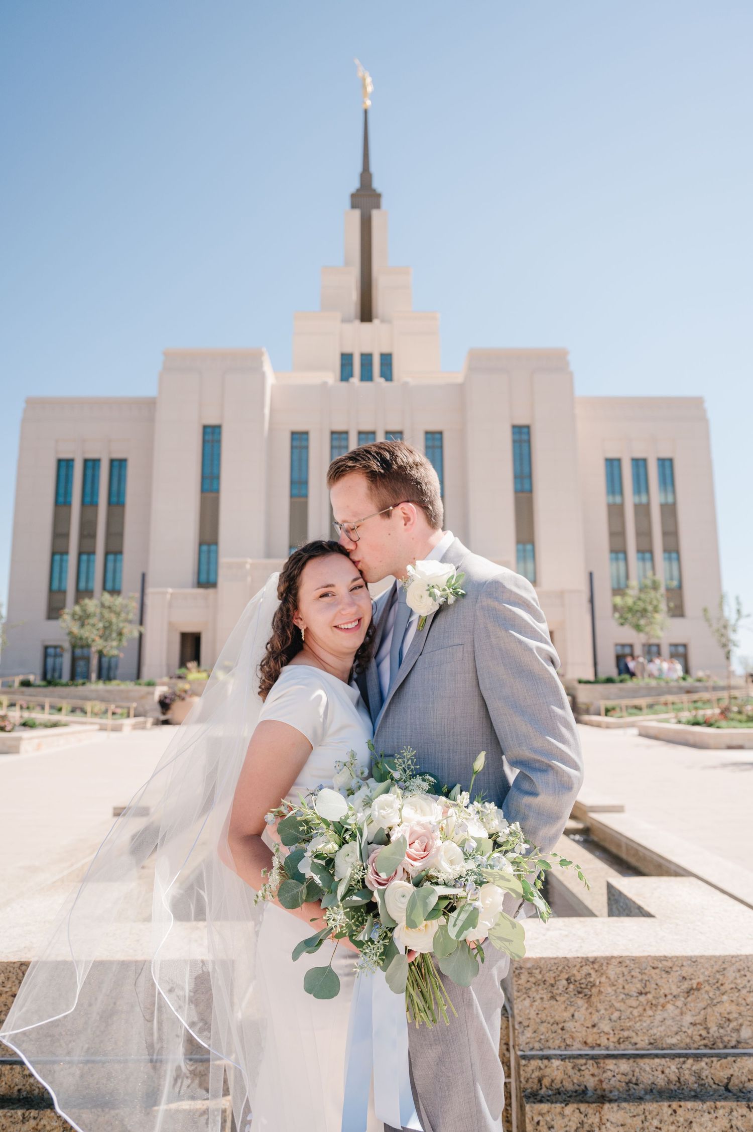 A couple embraces outside a Mormon temple on their wedding day with a beautiful floral bouquet.