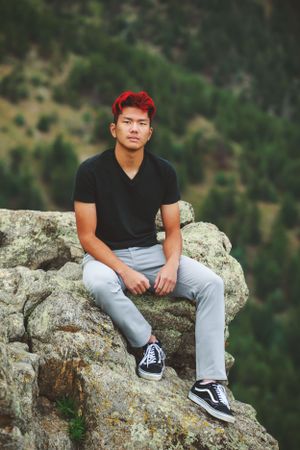 Person with vibrant red hair sitting on mountain ledge wearing black v-neck and light jeans.