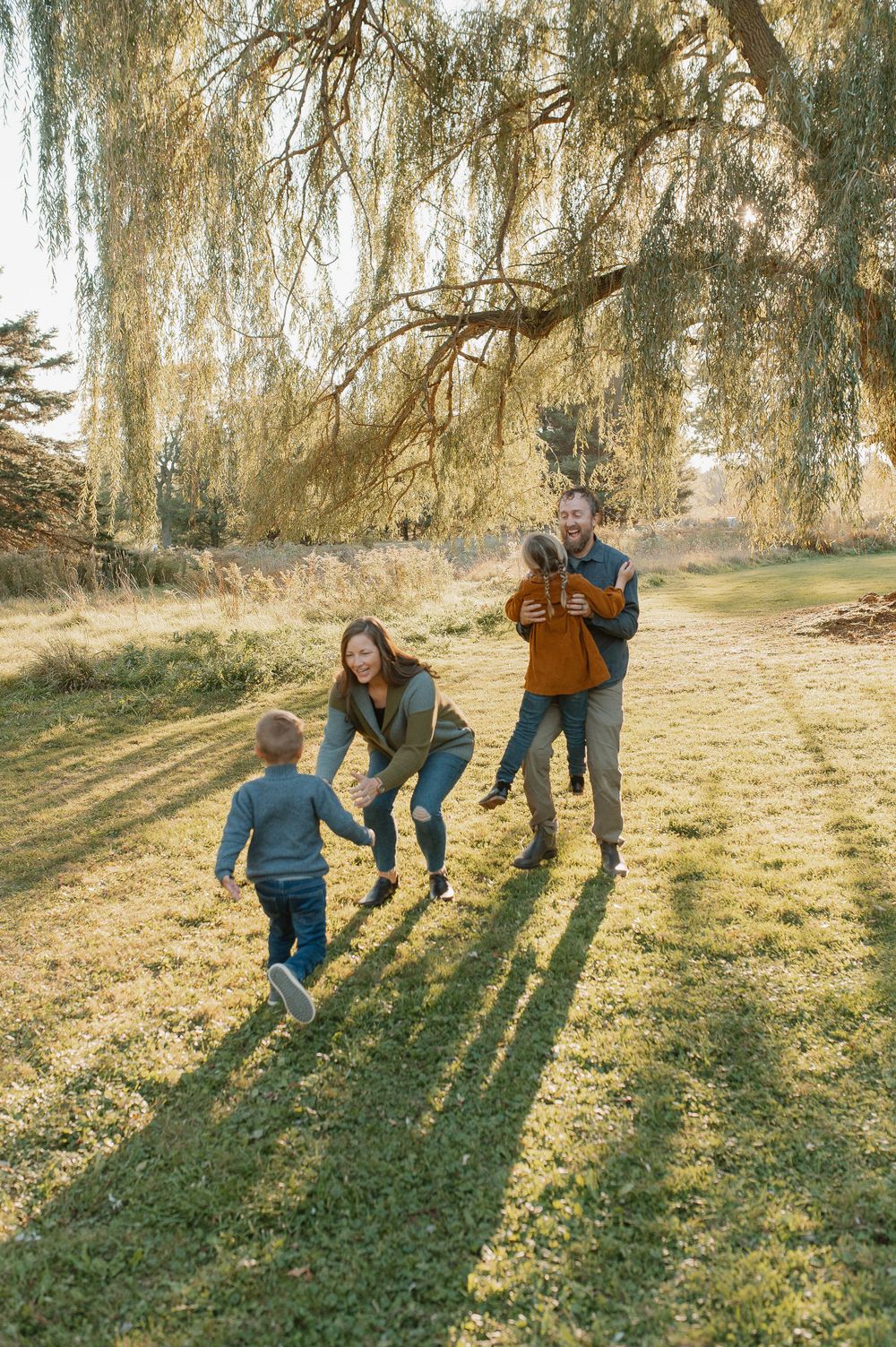Family plays together in a sunlit garden under drooping willow branches casting long afternoon shadows.