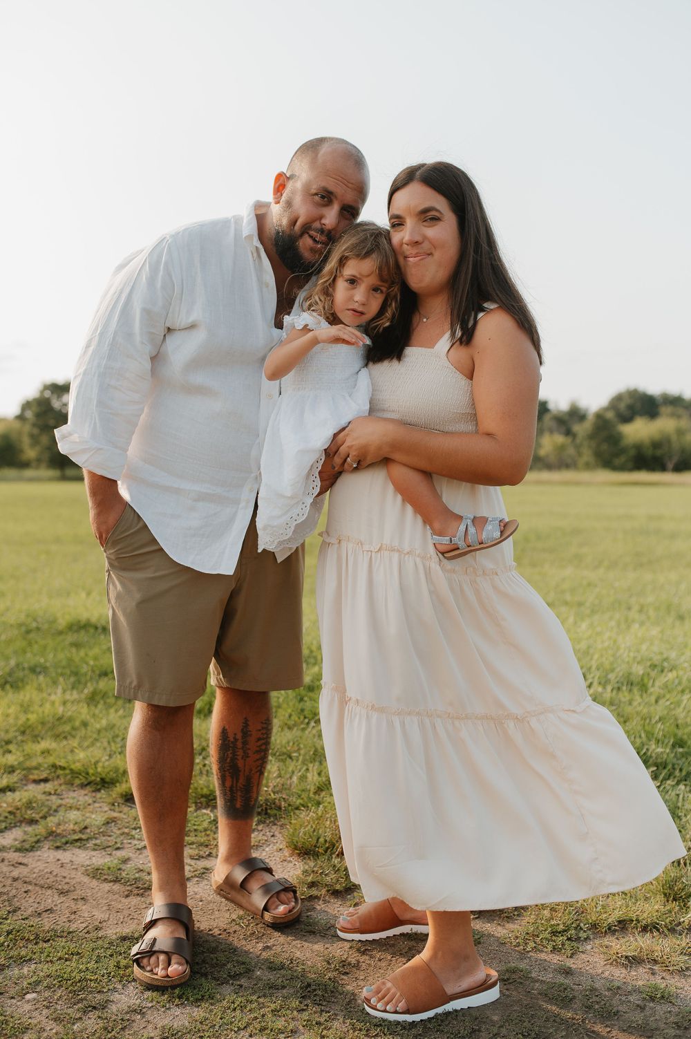 Family of three posing together in a summer field wearing light colored clothing.
