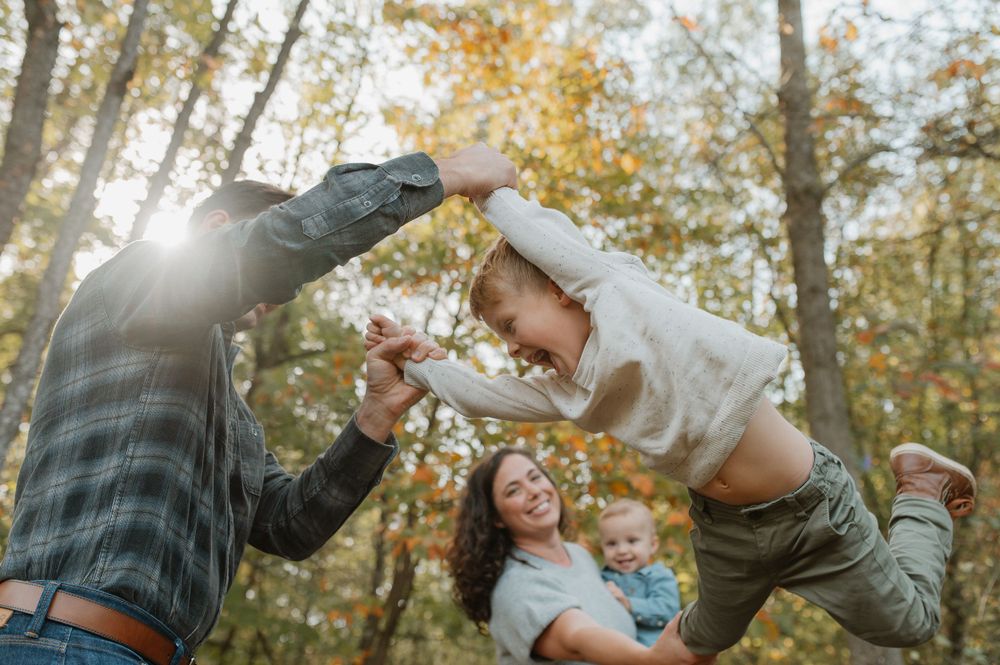 Family playing and lifting together in autumn forest with sunlight streaming through trees.