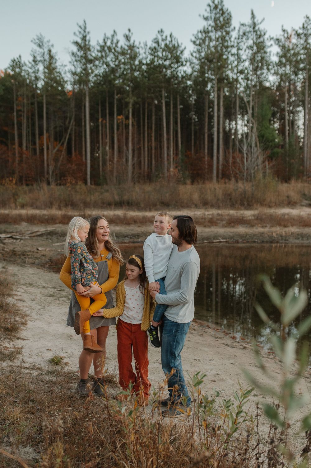 A group of people sharing laughter by a lake with tall pine trees in the background.