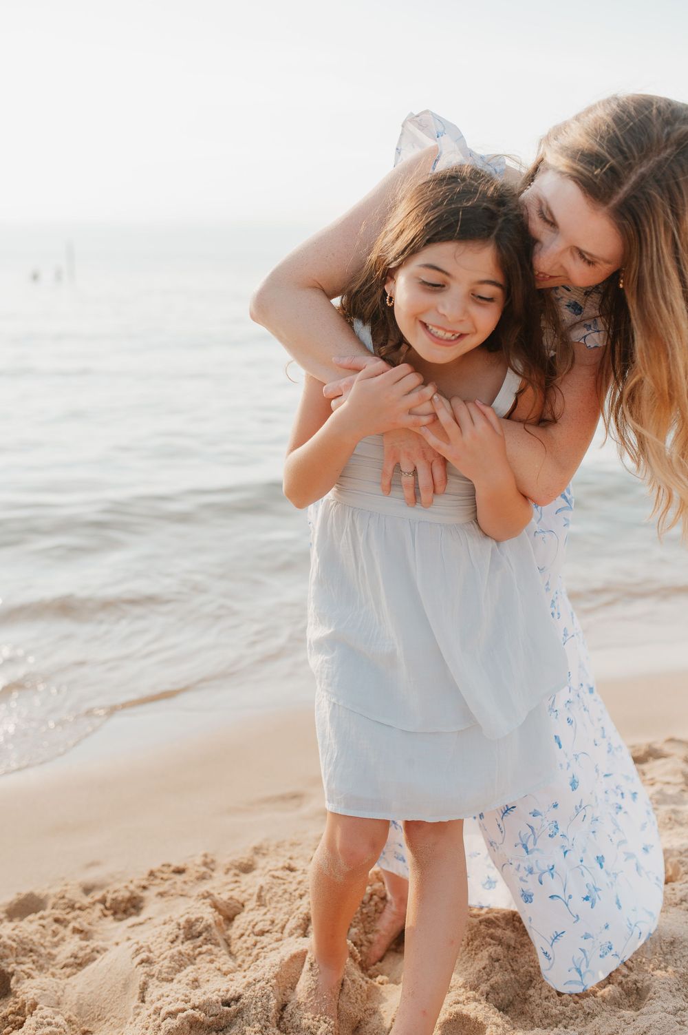 Two people share a joyful embrace on a sunny beach with waves in the background.