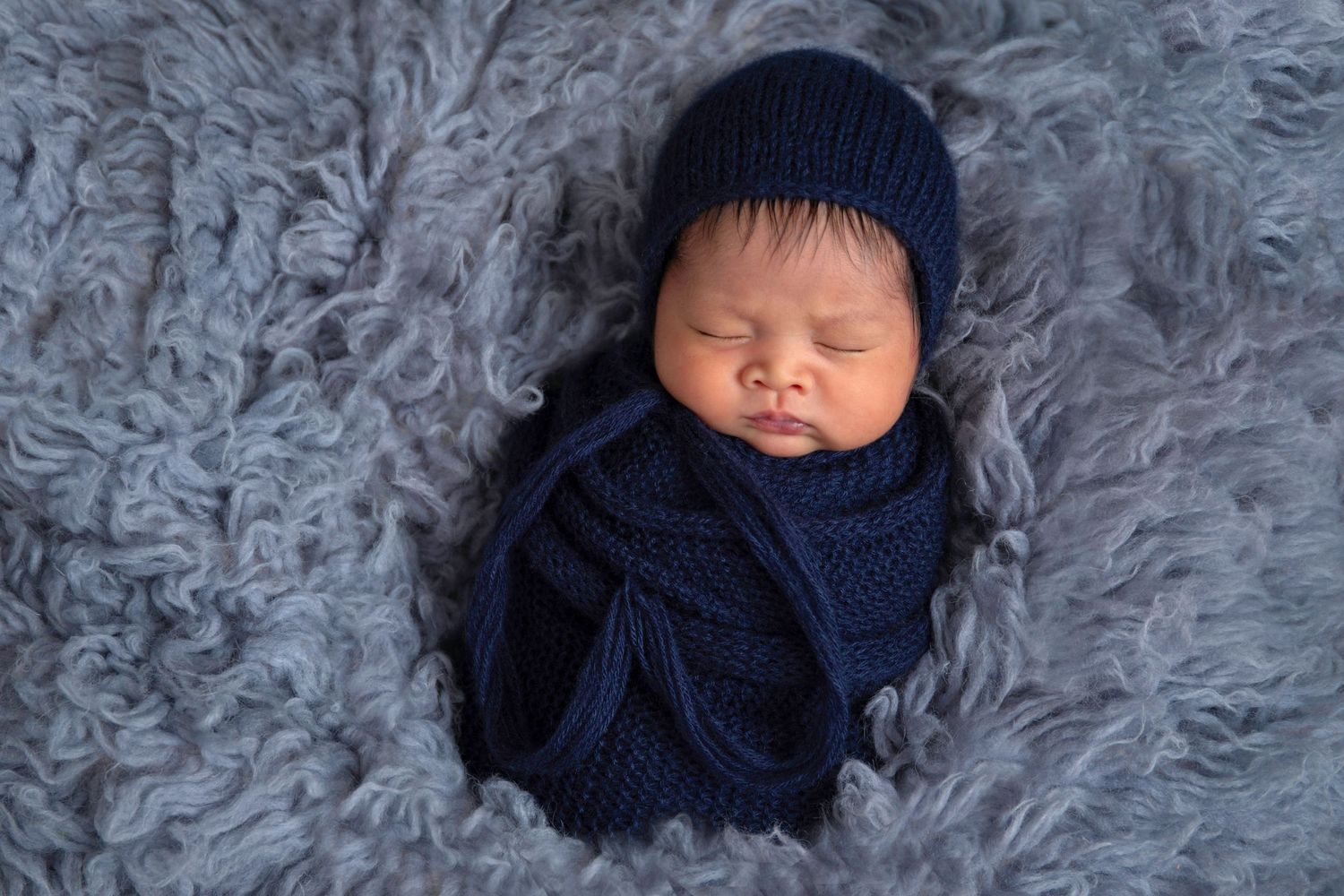 Peaceful sleeping newborn baby wrapped in navy knit blanket and beanie on soft gray fur background.