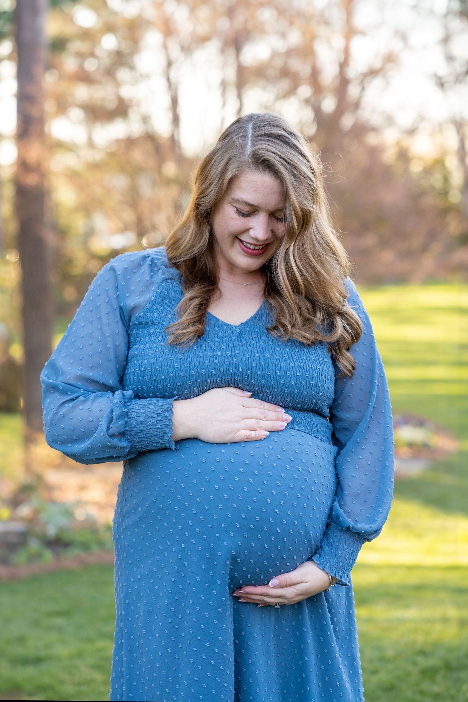 A woman in a blue dress stands in WRAL Azalea Garden holding her baby bump.