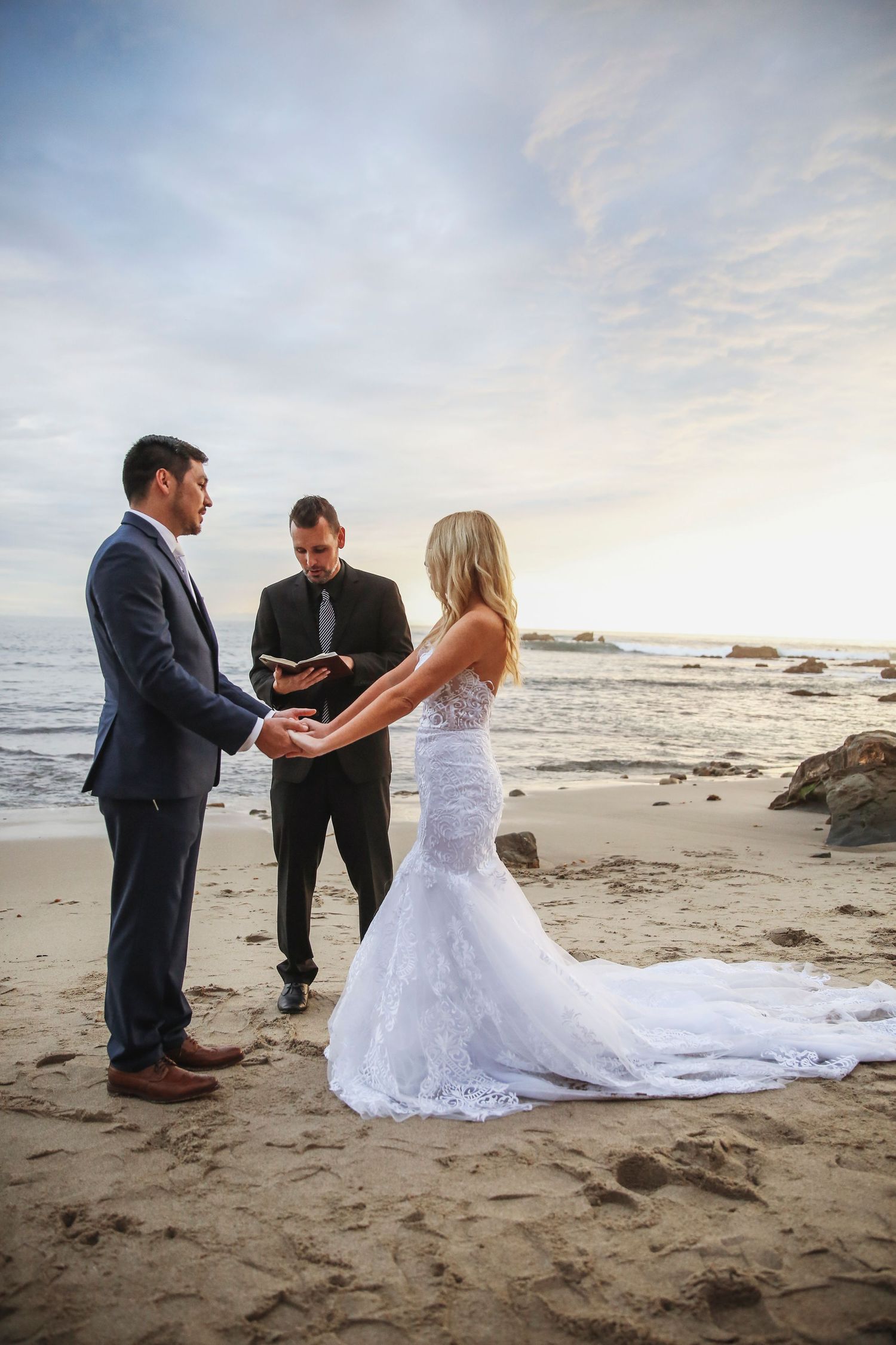 A small wedding ceremony takes place on a rocky beach at sunset with waves crashing in the background.