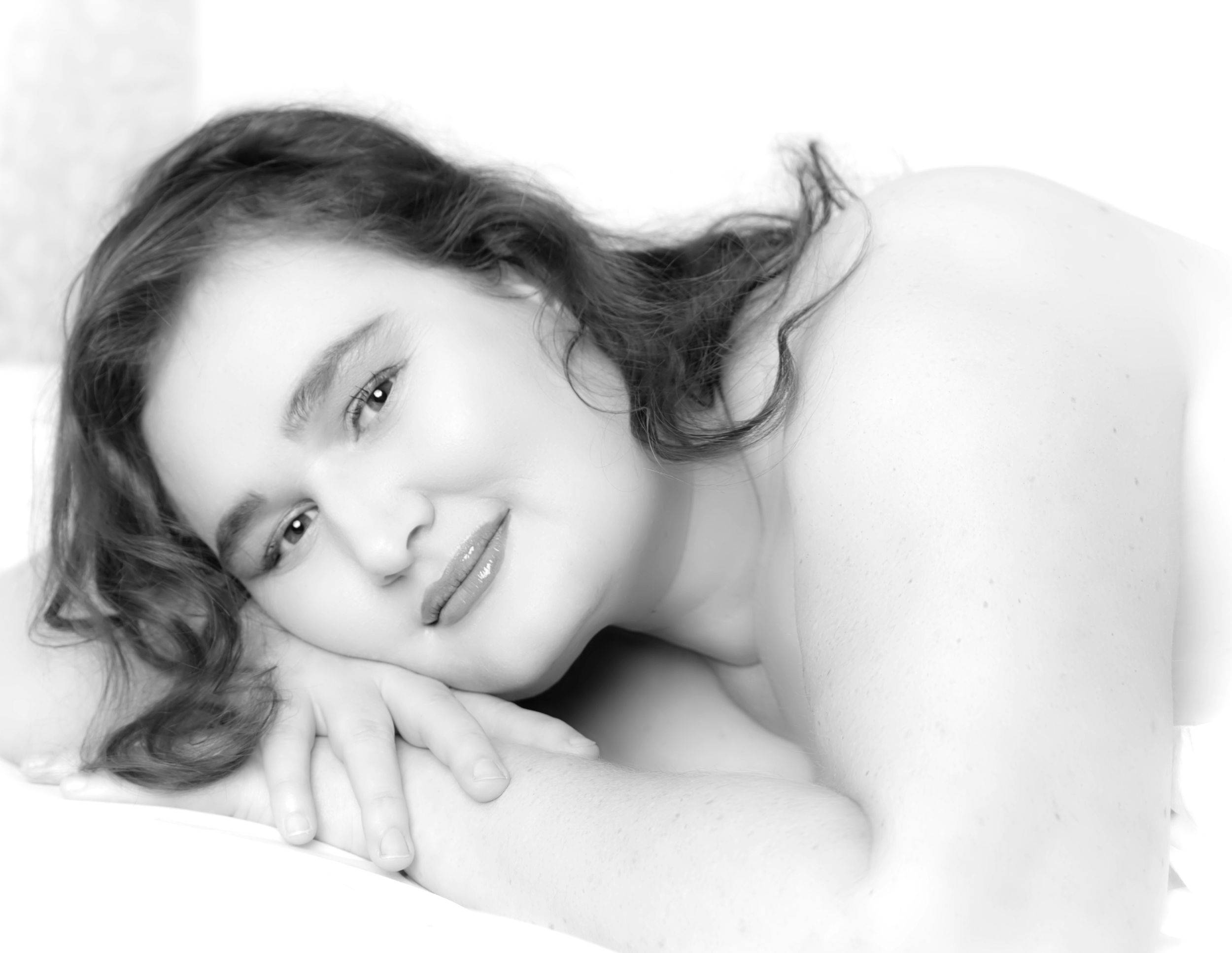 Black and white boudoir portrait in soft lighting showing gentle expression against white bedding.