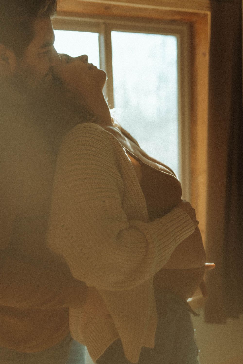 Side profile of a couple embracing near a window with soft ambient lighting.