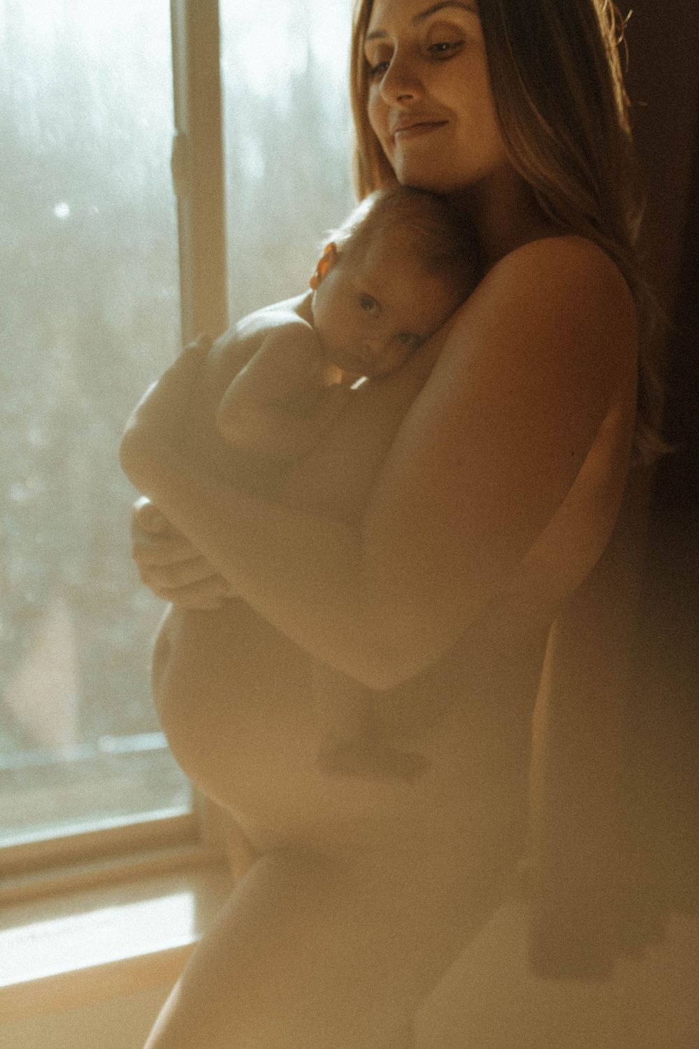 An intimate maternity portrait backlit by window light showing a nurturing moment.