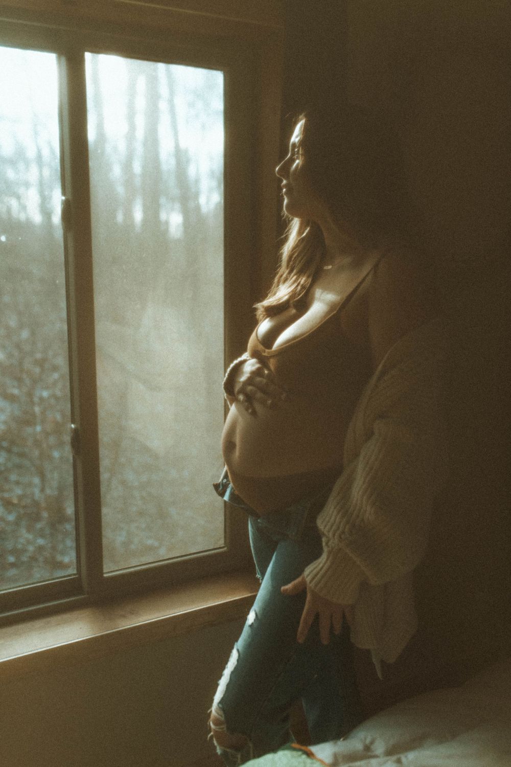 Silhouetted figure in cozy sweater standing by window with forest view beyond.
