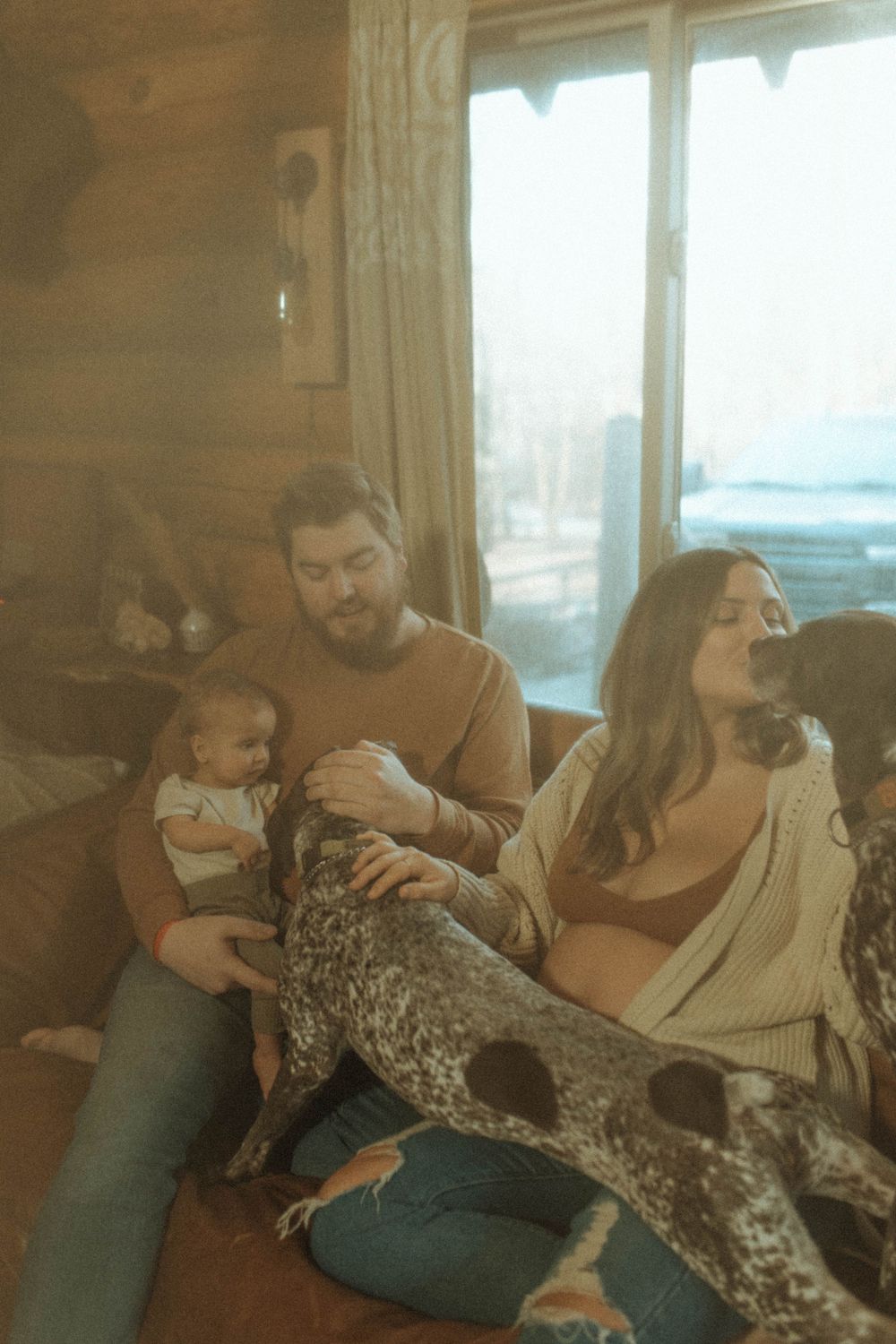 Family relaxing on couch with baby and dog in rustic cabin setting.