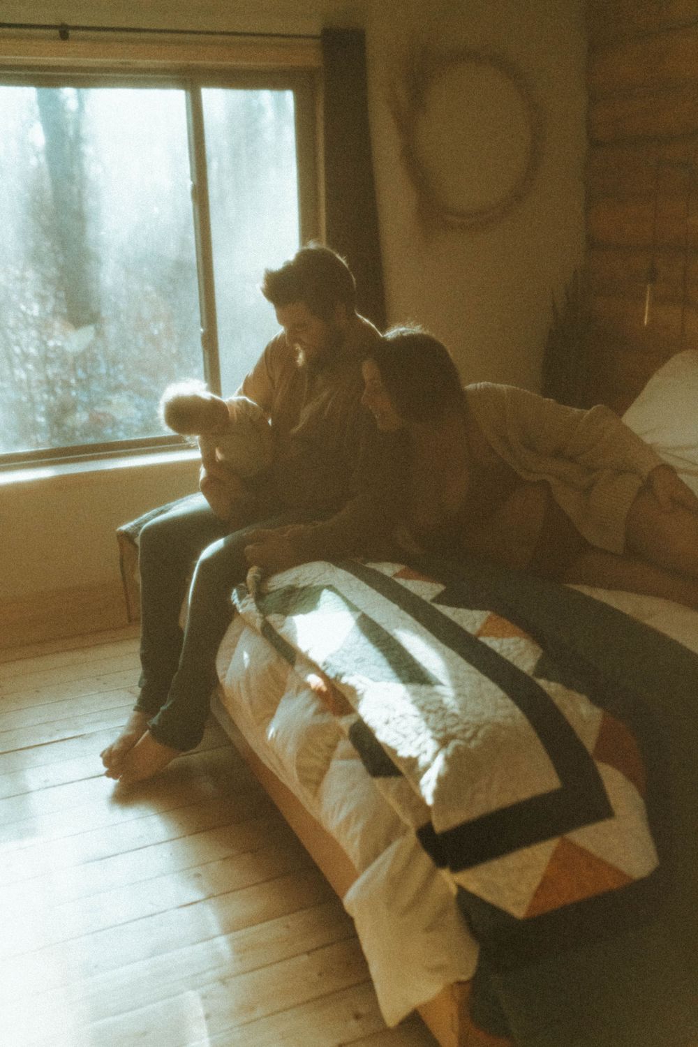 Soft natural window light illuminates figures sitting on a bed with a patterned quilt.