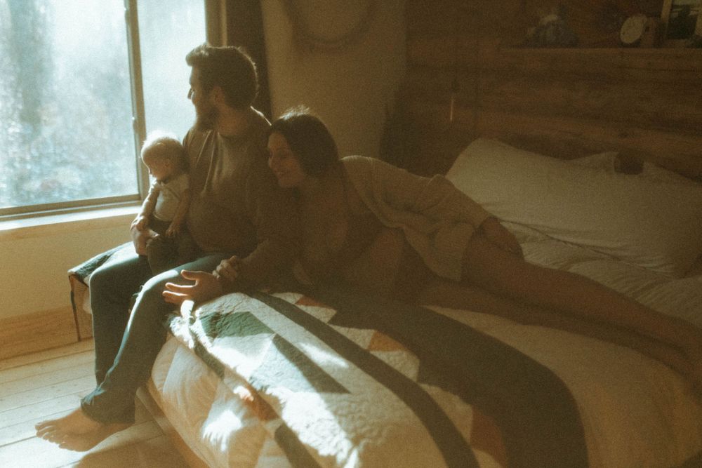 A dreamy film-style photo of a couple sitting by a bright window on a quilt-covered bed.