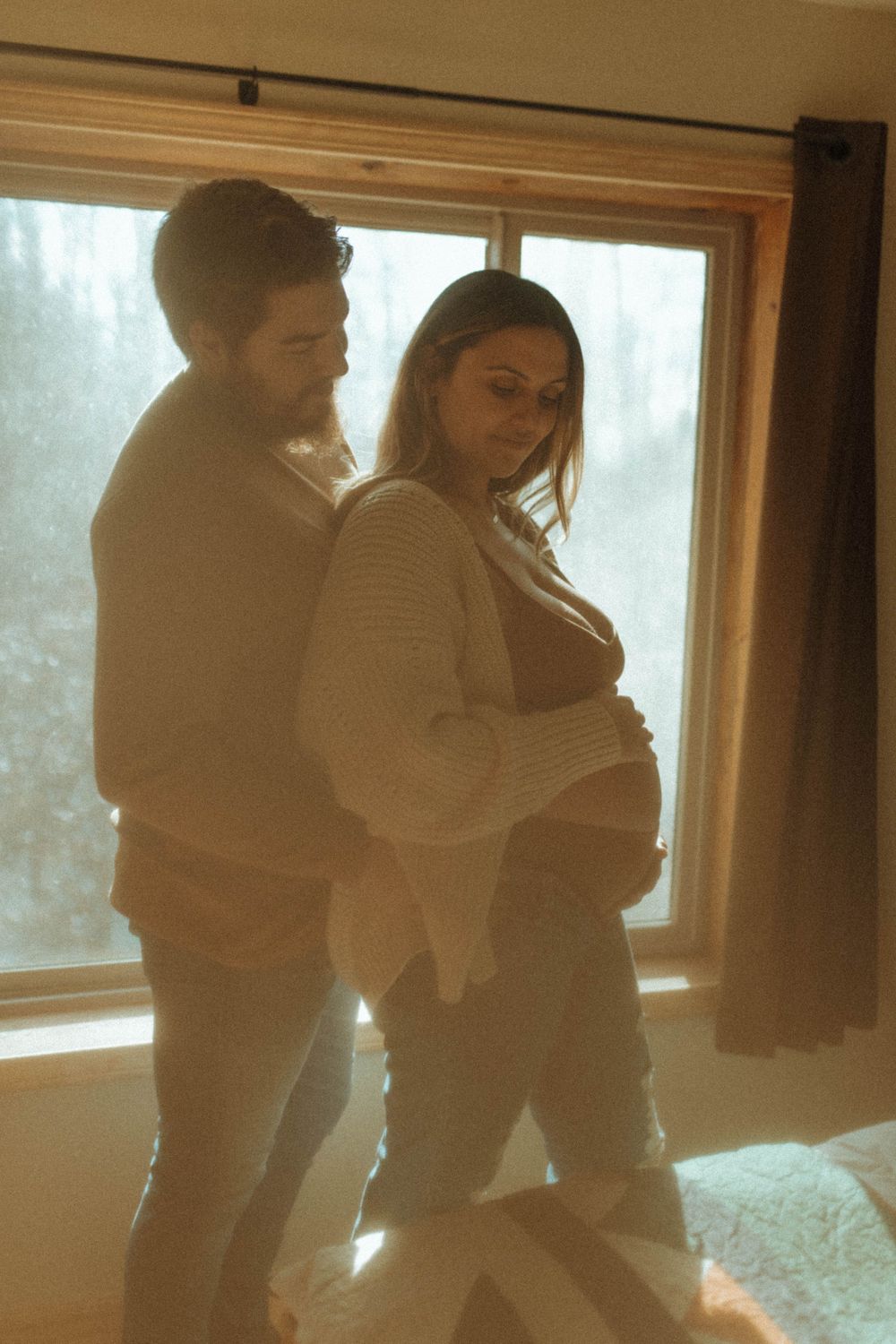A couple stands together in front of a window in a cozy interior setting.