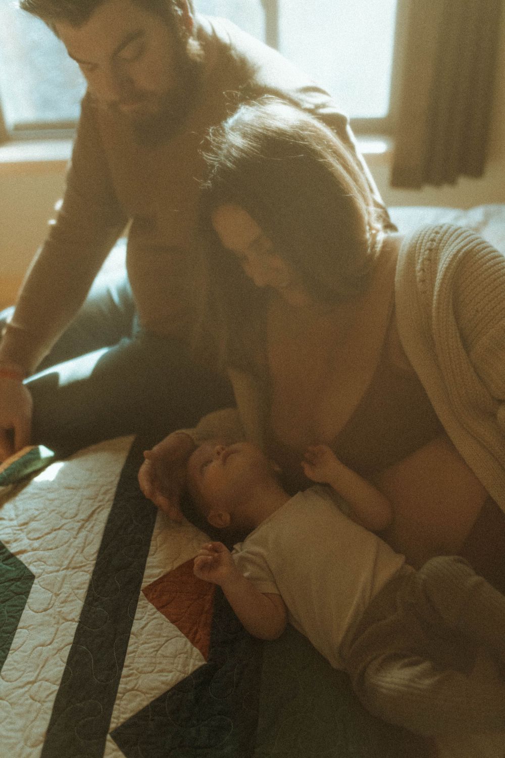 A tender family moment with an infant on a patterned quilt in warm lighting.