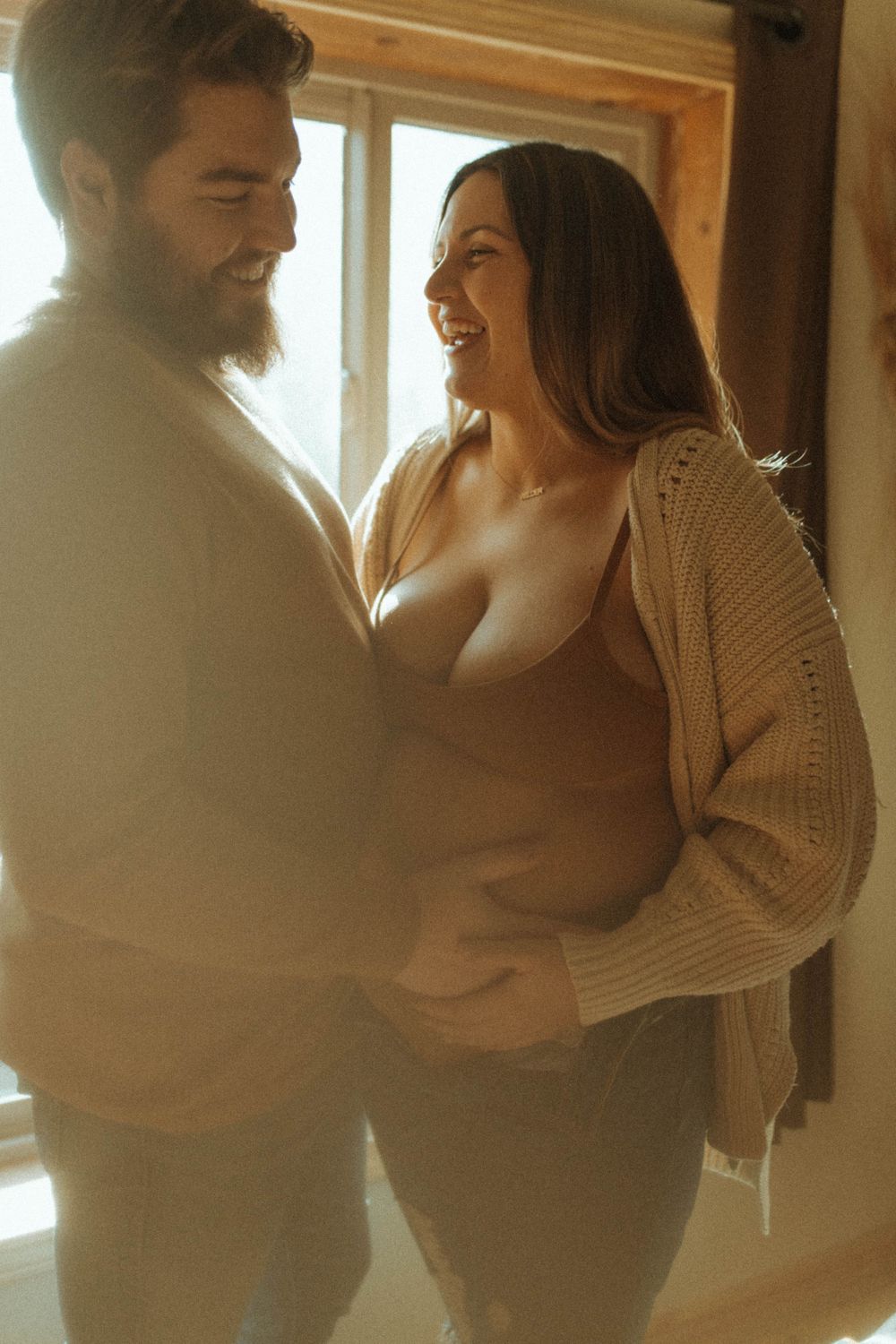 A couple shares an intimate moment together by a window in warm lighting.