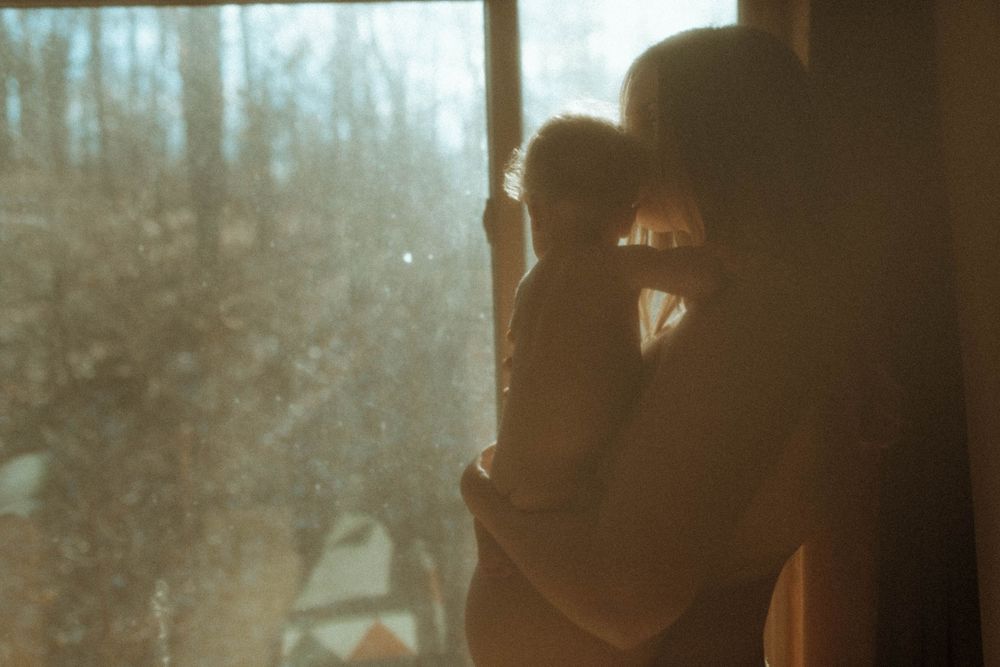 Silhouetted figures stand by a window in warm sunlight creating an intimate maternal moment.