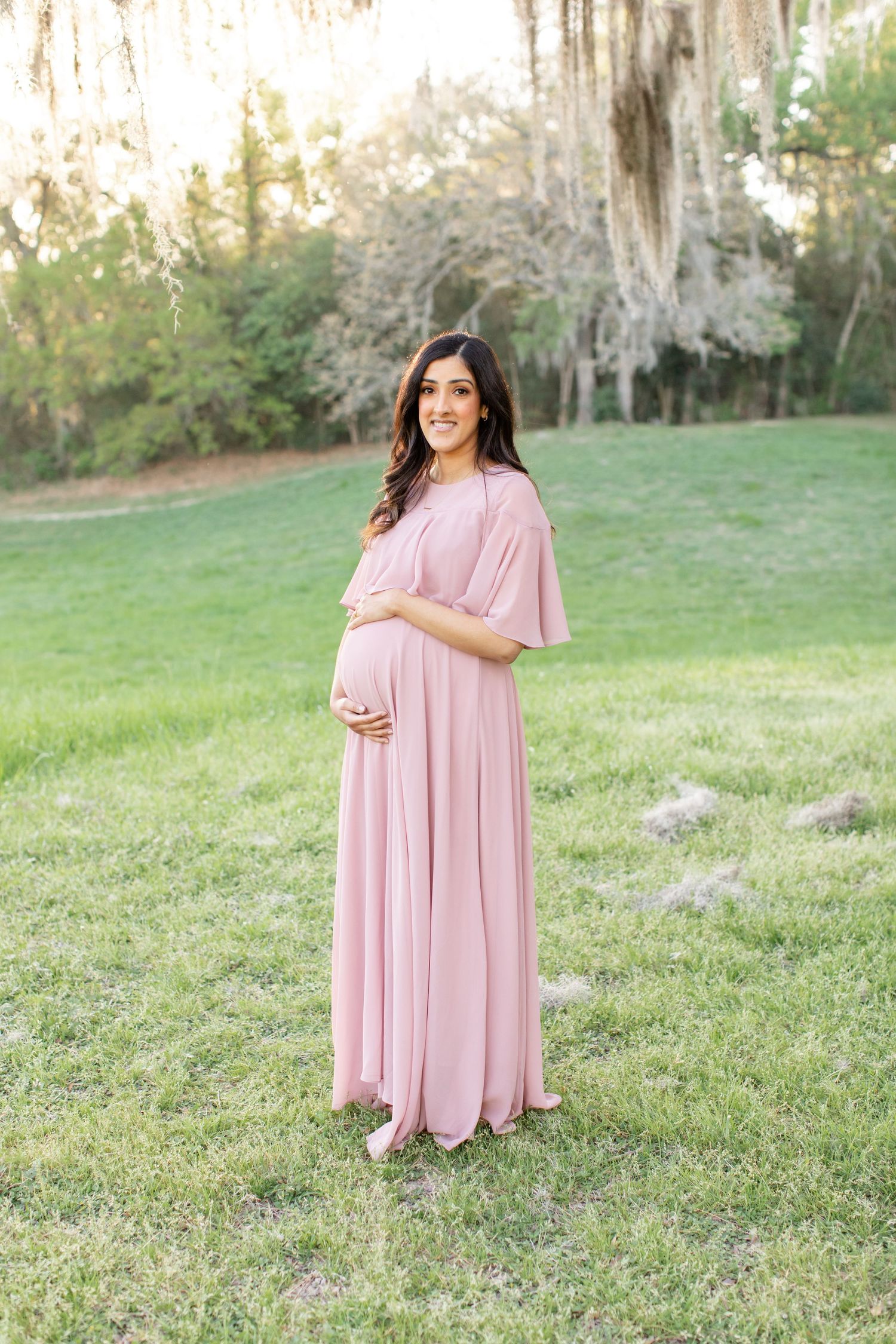 A radiant maternity portrait features a flowing pink dress in a mossy outdoor setting.