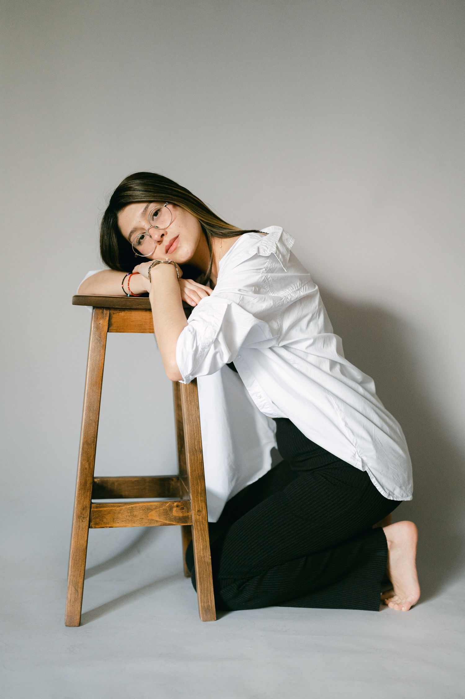 Portrait of person in white shirt leaning on wooden stool against neutral background.