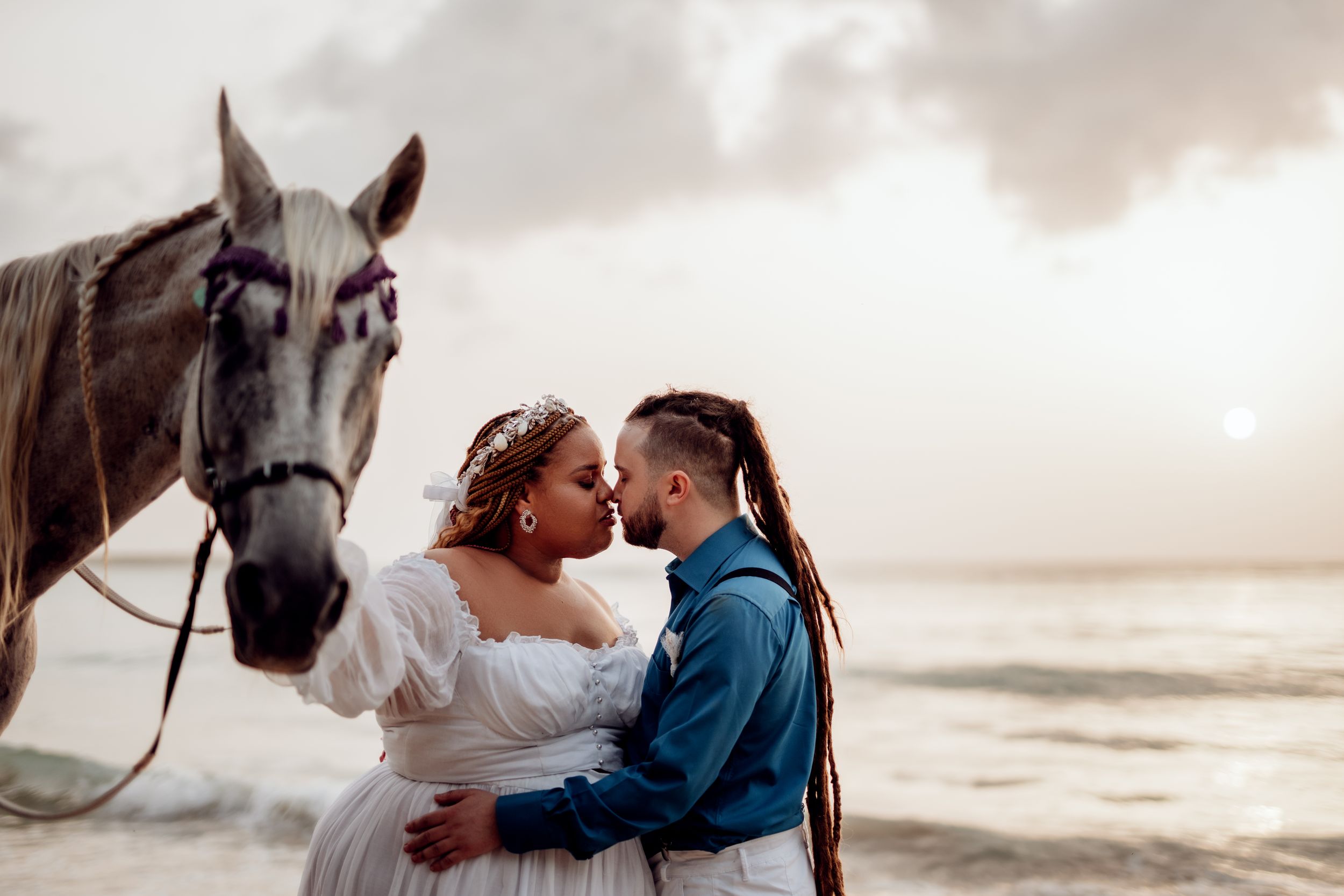 A romantic beach wedding scene with a horse standing beside the couple at sunset.