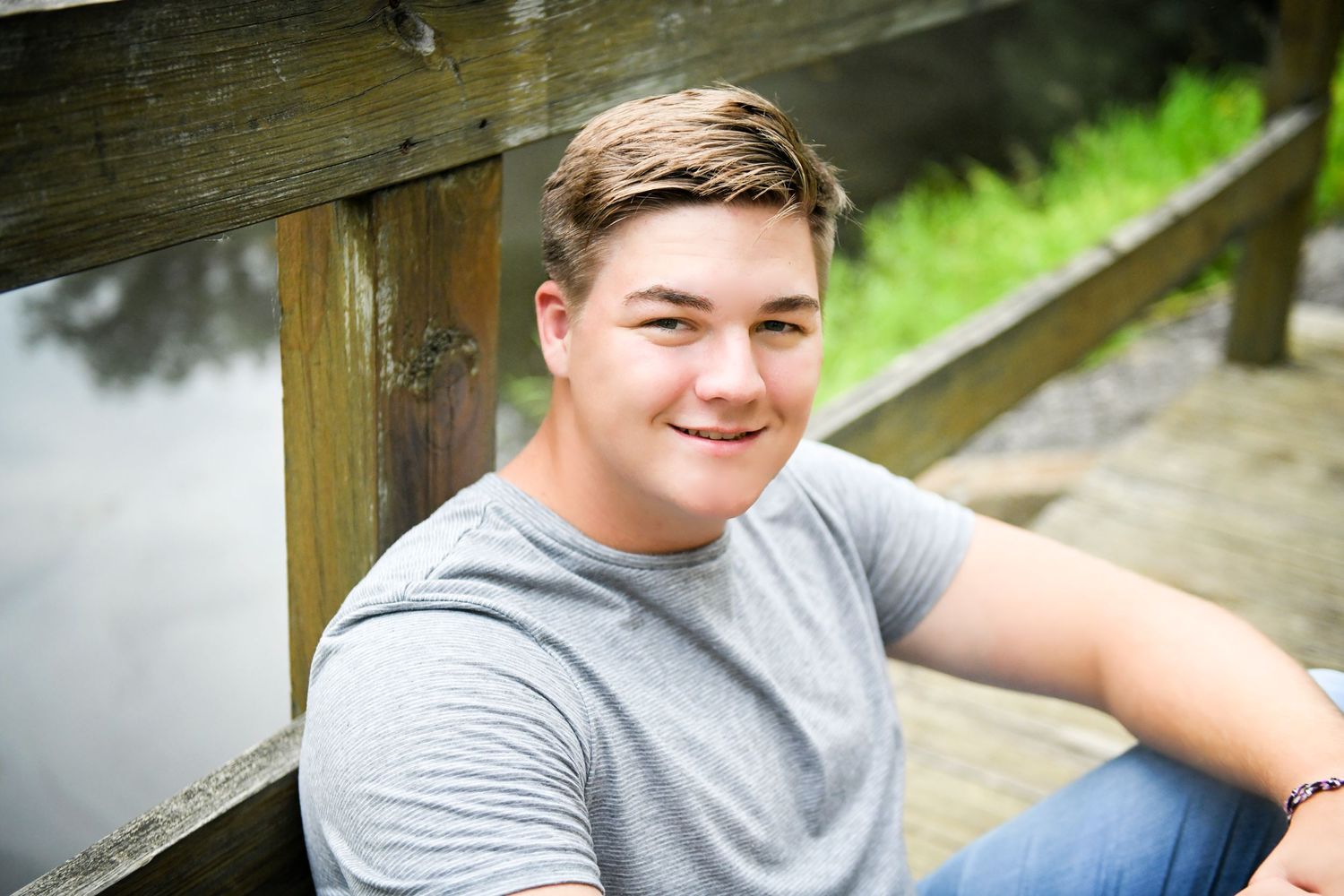 Smiling young man in gray t-shirt sitting on wooden dock near water outdoors.