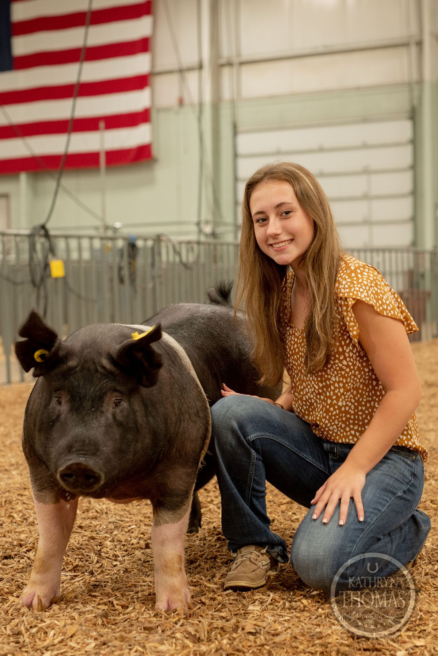 Show pig with distinctive black and white coloring stands in sawdust pen with American flag in background.