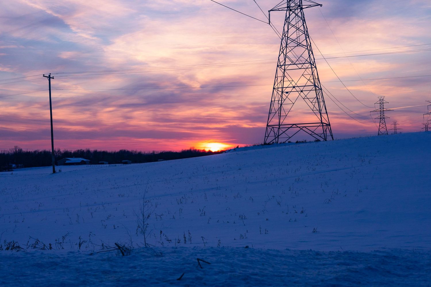Electrical transmission tower silhouetted against a pink and purple winter sunset over snowy terrain.