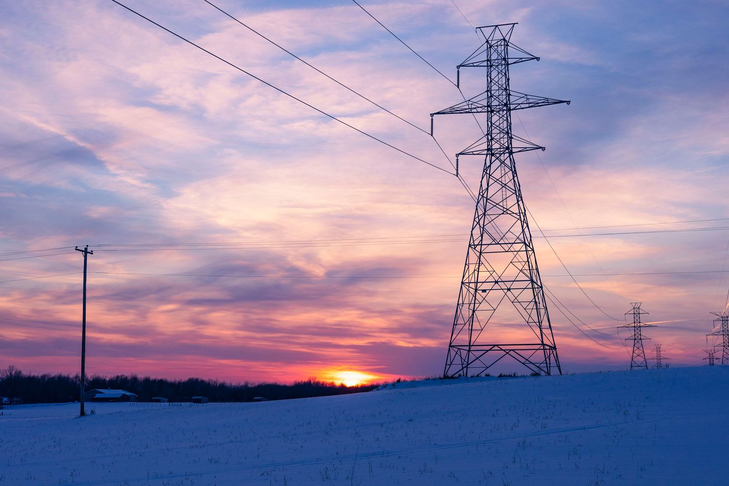 Power lines and tower frame a vibrant sunset over a snow-covered landscape.