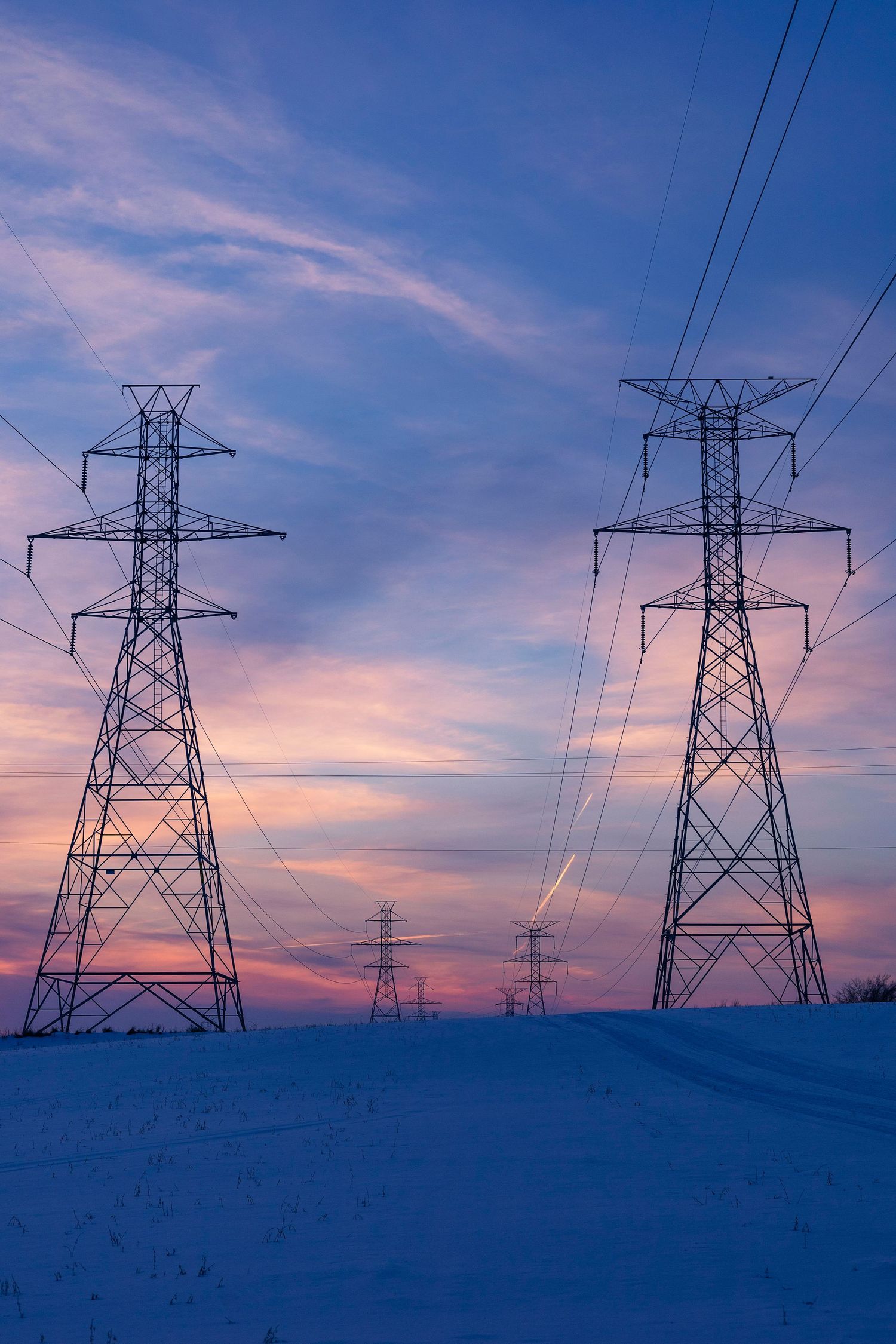 Multiple electrical towers stand against a colorful winter sunset sky with snow below.