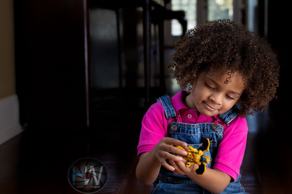Child in denim overalls playing with yellow toy car indoors.