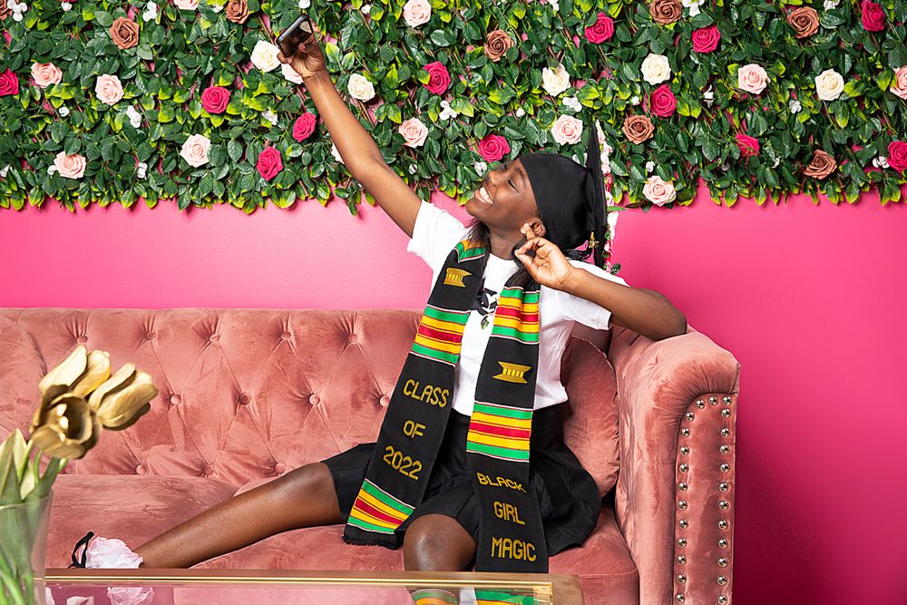Graduate in cap and gown celebrating against pink wall with floral backdrop.