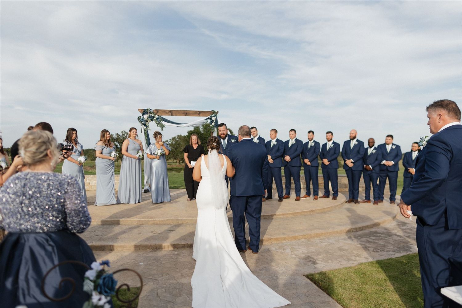 Outdoor wedding ceremony with bridal party in navy and light blue on stone patio under blue skies.