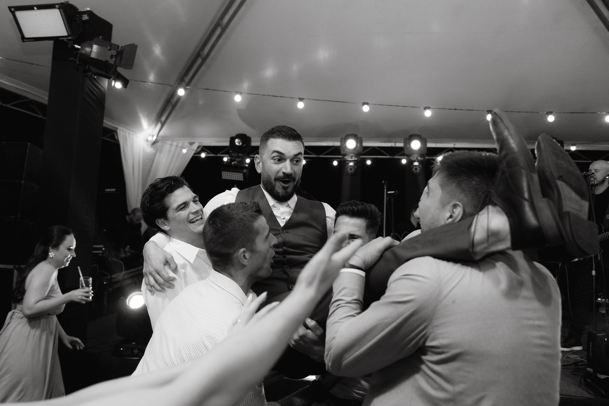 Black and white photo of wedding guests dancing and celebrating under tent lighting.