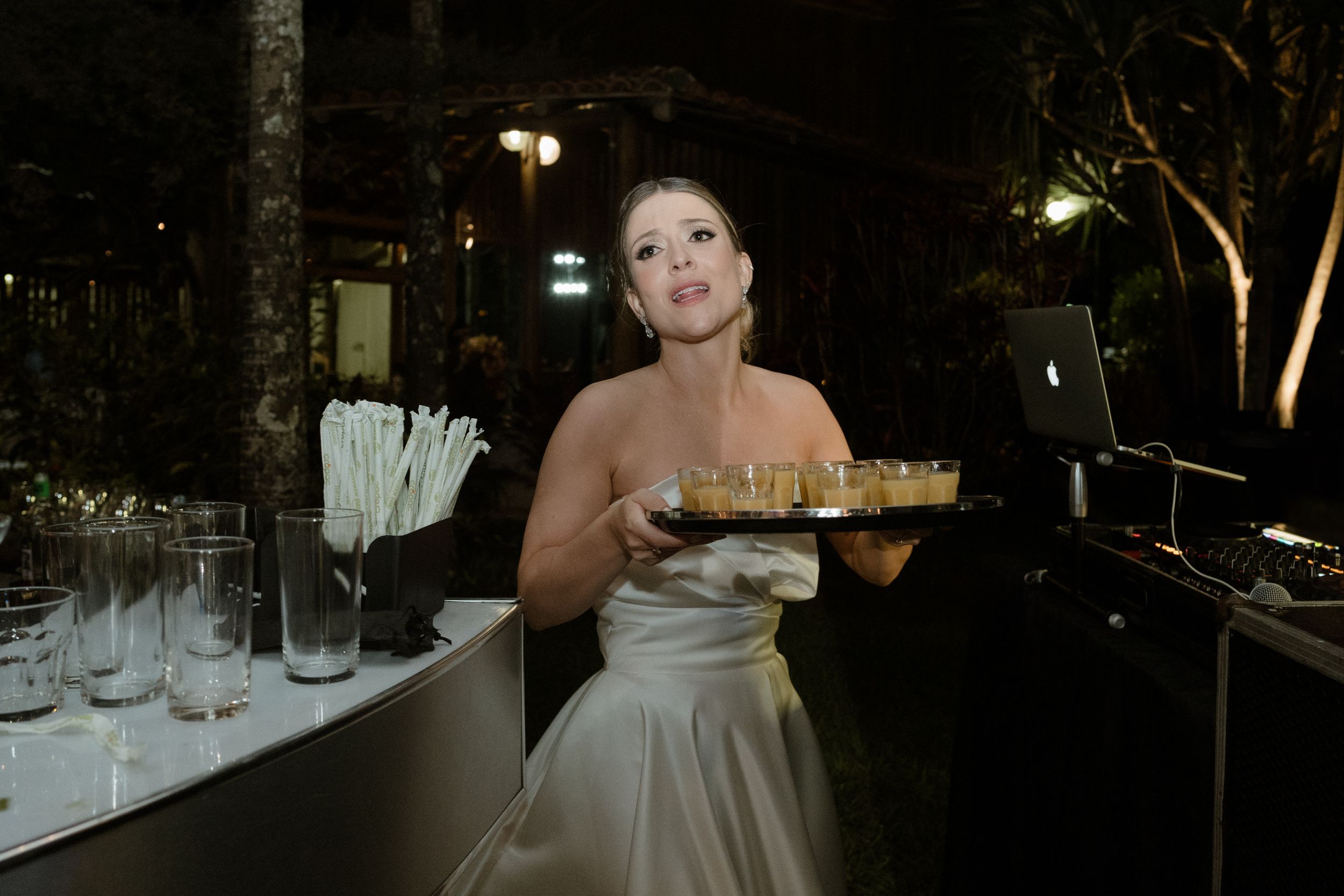 Bride in white dress holding food plate during nighttime outdoor reception.