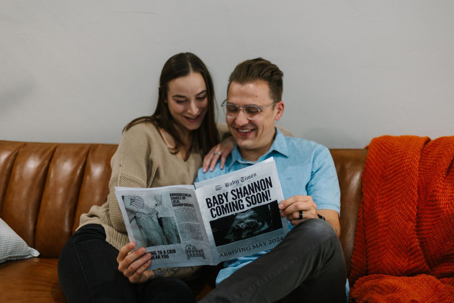 A couple reads a newspaper together while relaxing on a leather couch with an orange throw blanket.