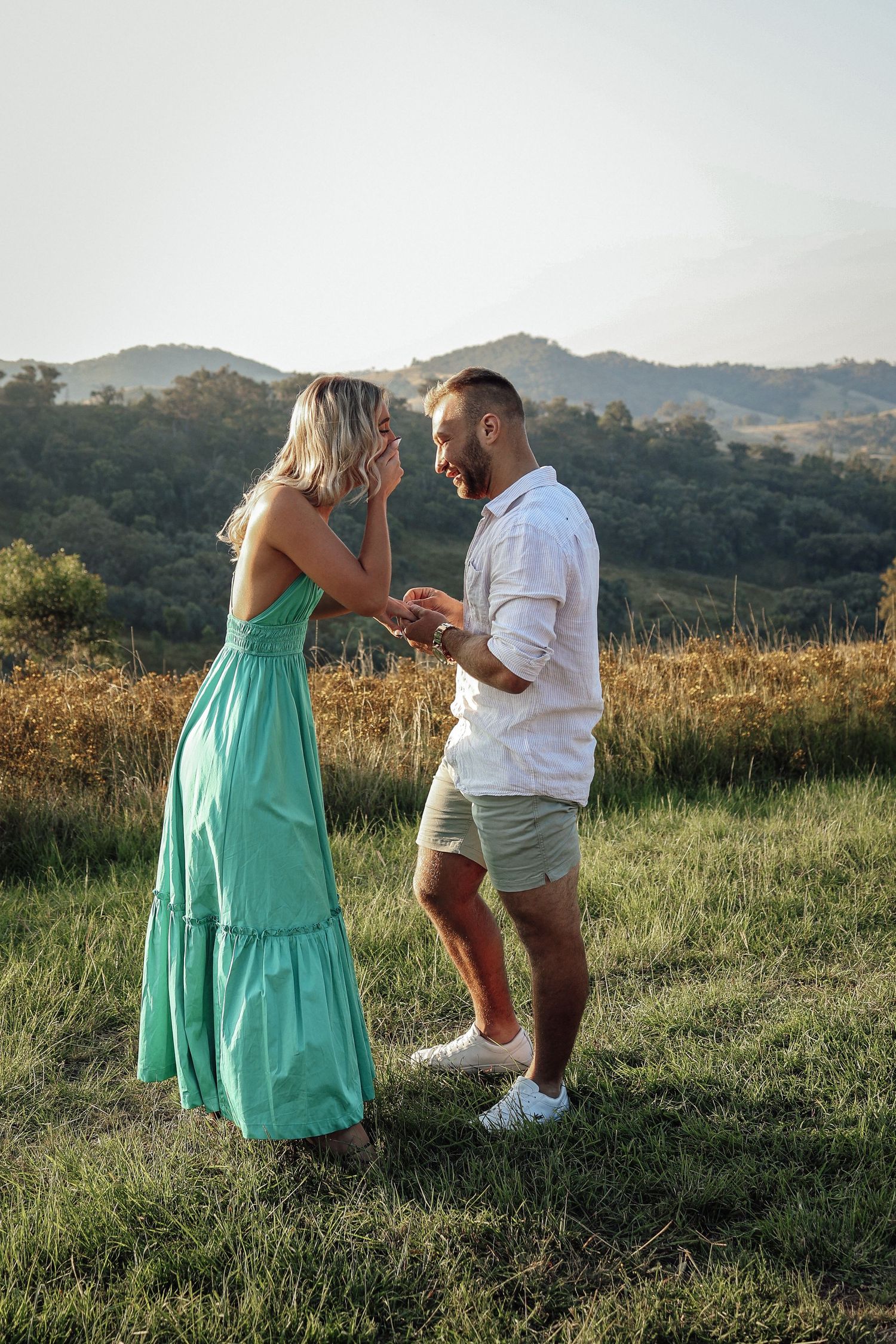 A couple shares an emotional moment in a grassy field with mountains behind them.