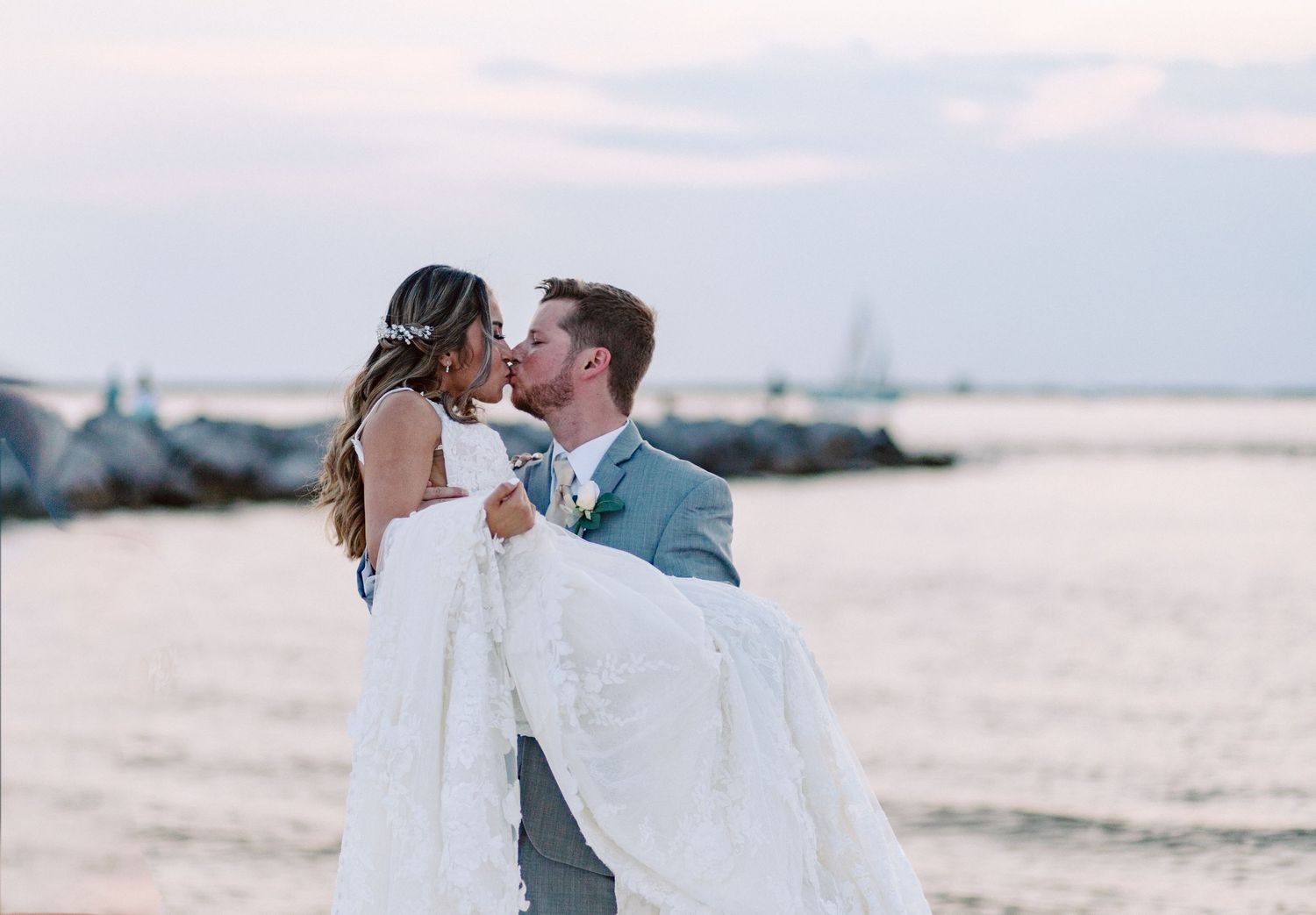 A romantic beachside wedding couple embraces as the groom carries the bride in her flowing white dress at sunset.