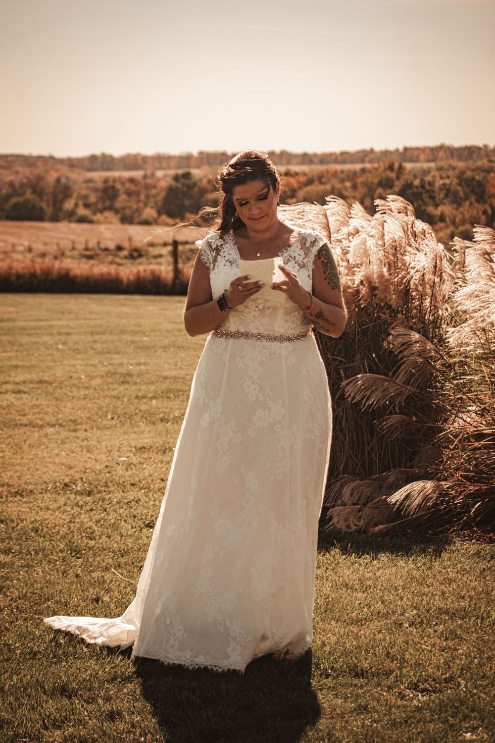 A bride stands in a flowing lace gown reading a letter in front of rustic pampas grass.