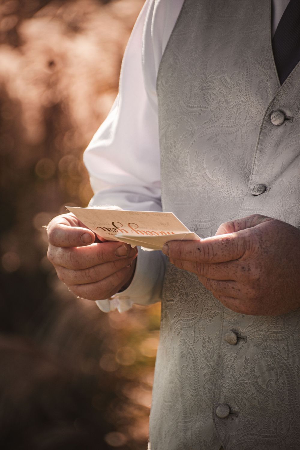 Close-up of person in formal vest holding a handwritten note in golden sunlight.