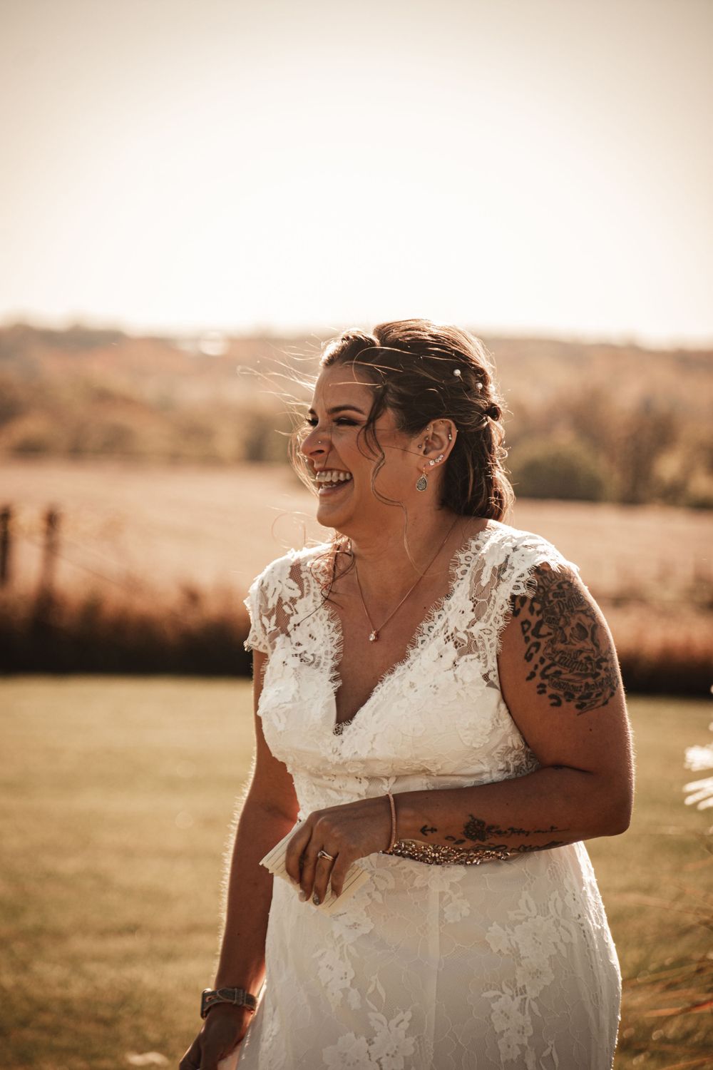 Candid moment of bride laughing in lace dress with visible tattoos in golden outdoor setting.